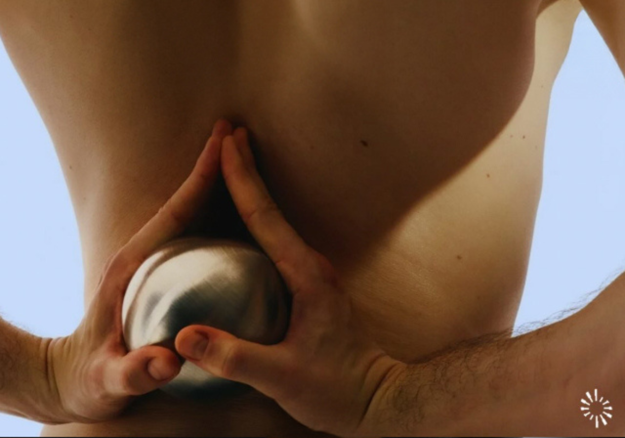 Zbliżenie dolnej części pleców mężczyzny trzymającego przy kręgosłupie metalową kulę do masażu. Dłonie dociskają kulę po obu stronach pleców, sugerując automasaż lub terapię punktów spustowych w okolicy lędźwiowej.